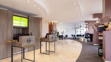A hotel reception area with a reception desk, a digital screen displaying information, and a seating area with tables and chairs.