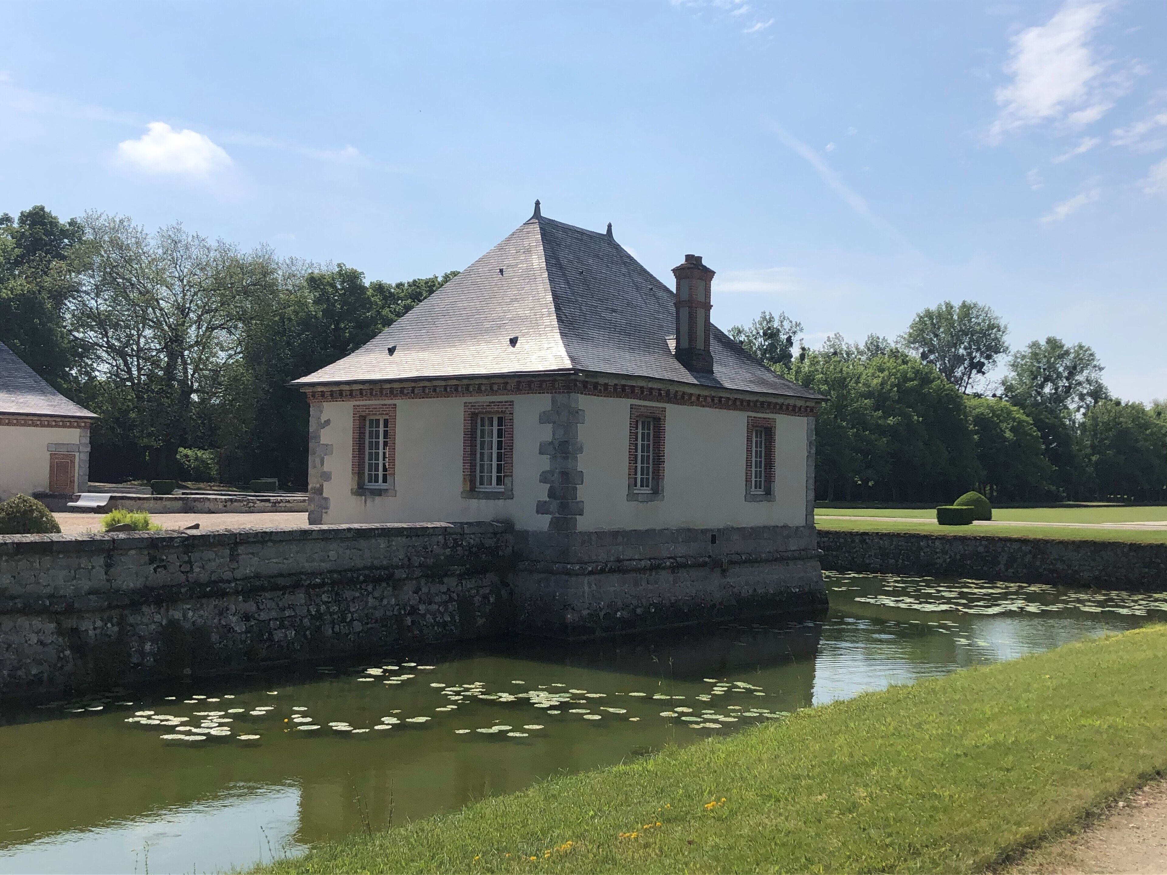 A small, stone building with a gabled roof, situated next to a calm water body with lily pads.