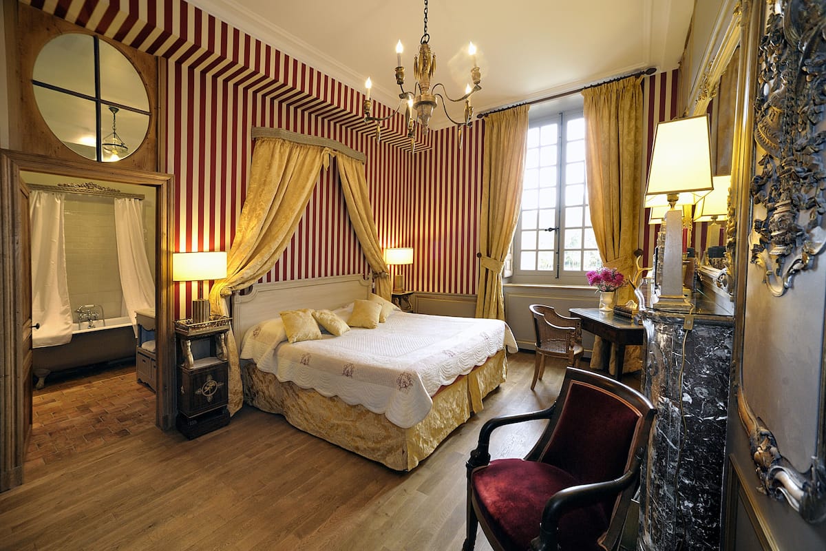 A hotel room with a large bed, a sitting area with a chair, a vanity with a mirror, and a chandelier.