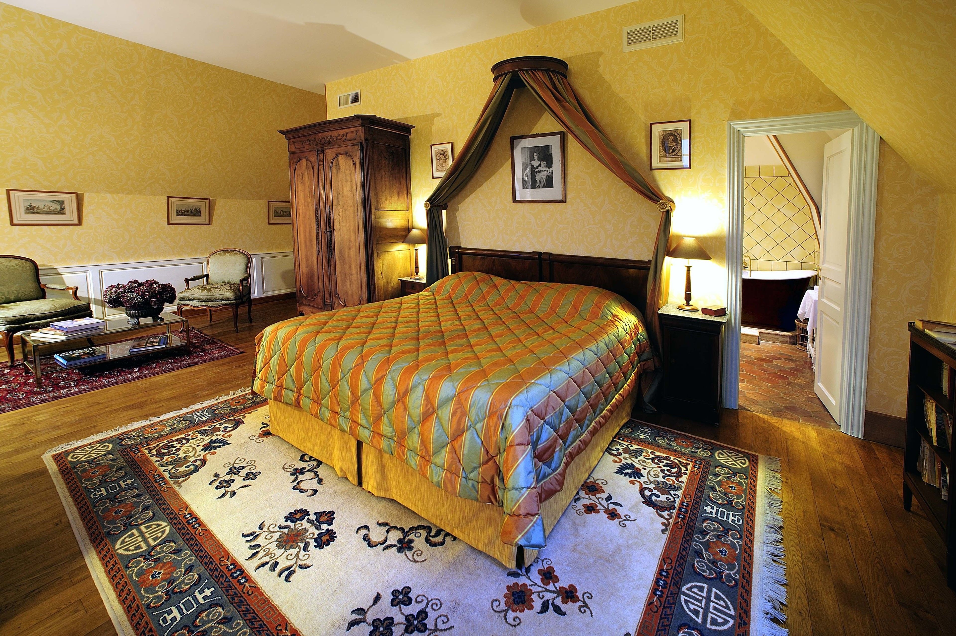 A bedroom with a large bed, a wooden wardrobe, a sitting area with a chair and table, and a staircase leading to another room.