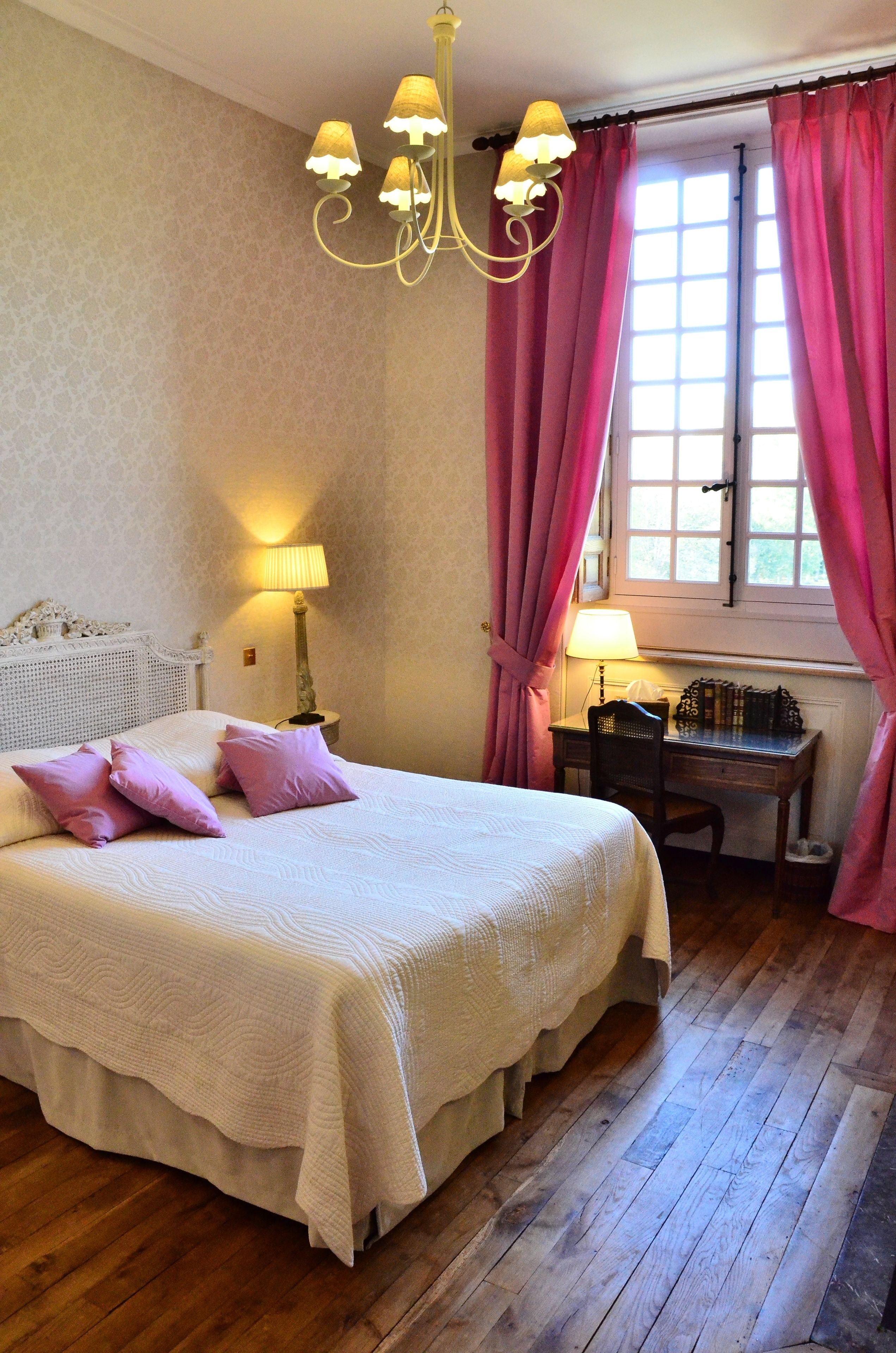 A bedroom with a large bed, a desk, a chair, a lamp, and a window with curtains.