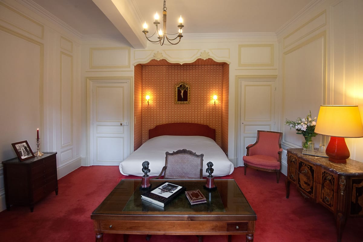 A hotel room with a large bed, a wooden coffee table, a red armchair, a nightstand, a lamp, and a framed picture on the nightstand.