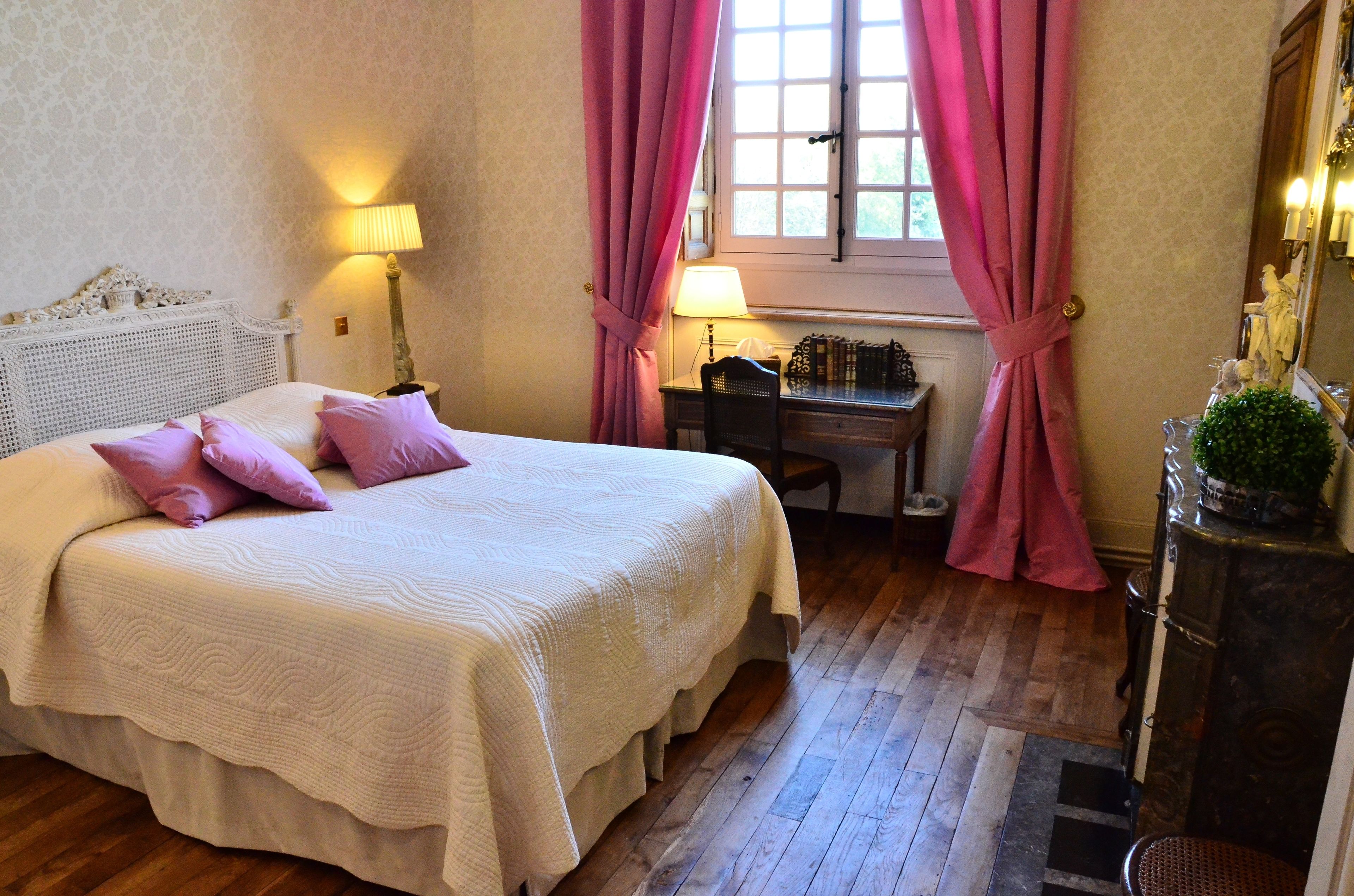 Una cama bien hecha con una colcha blanca y varias almohadas, un escritorio de madera con una lámpara y una ventana con cortinas rosadas.