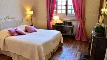 Una cama bien hecha con una colcha blanca y varias almohadas, un escritorio de madera con una lámpara y una ventana con cortinas rosadas.