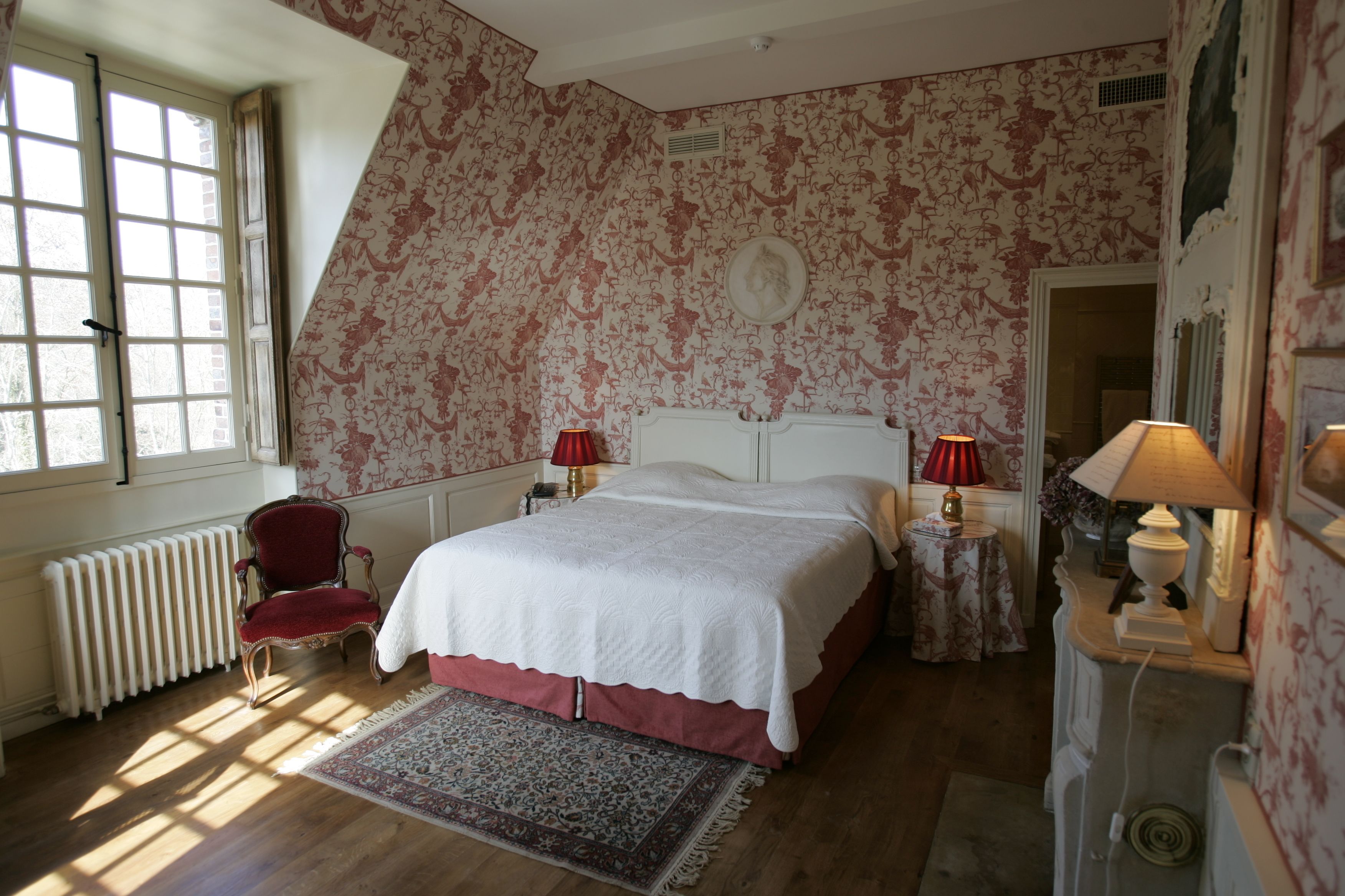 A bedroom with a large bed, a red chair, two bedside tables with lamps, a radiator, and a window with curtains.