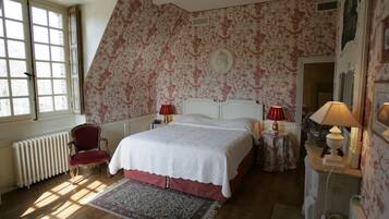 A bedroom with a large bed, a red chair, two bedside tables with lamps, a radiator, and a window with curtains.