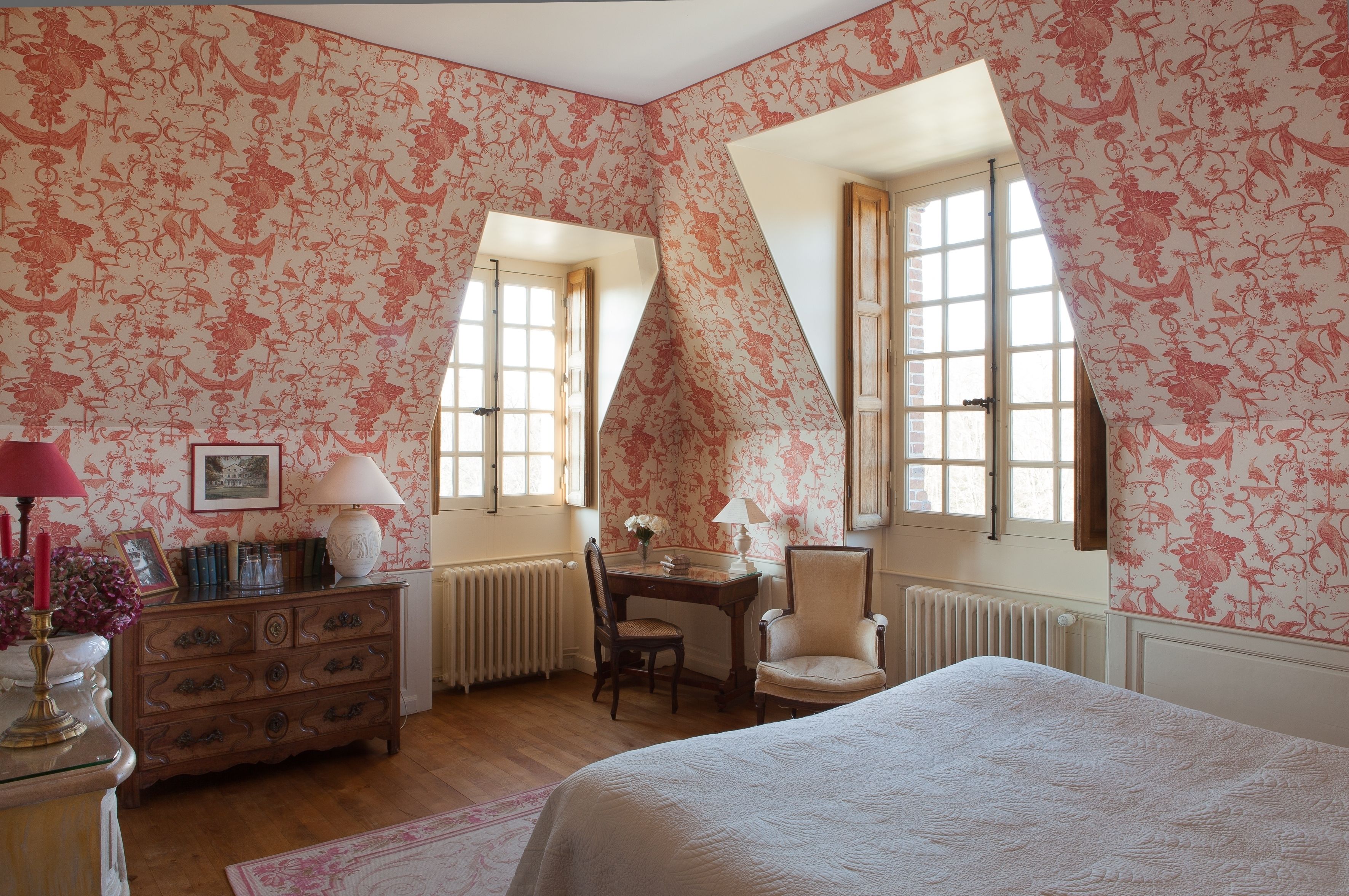 Un dormitorio con papel tapiz floral, un aparador de madera, un escritorio con silla, una cama y dos ventanas con marcos blancos.