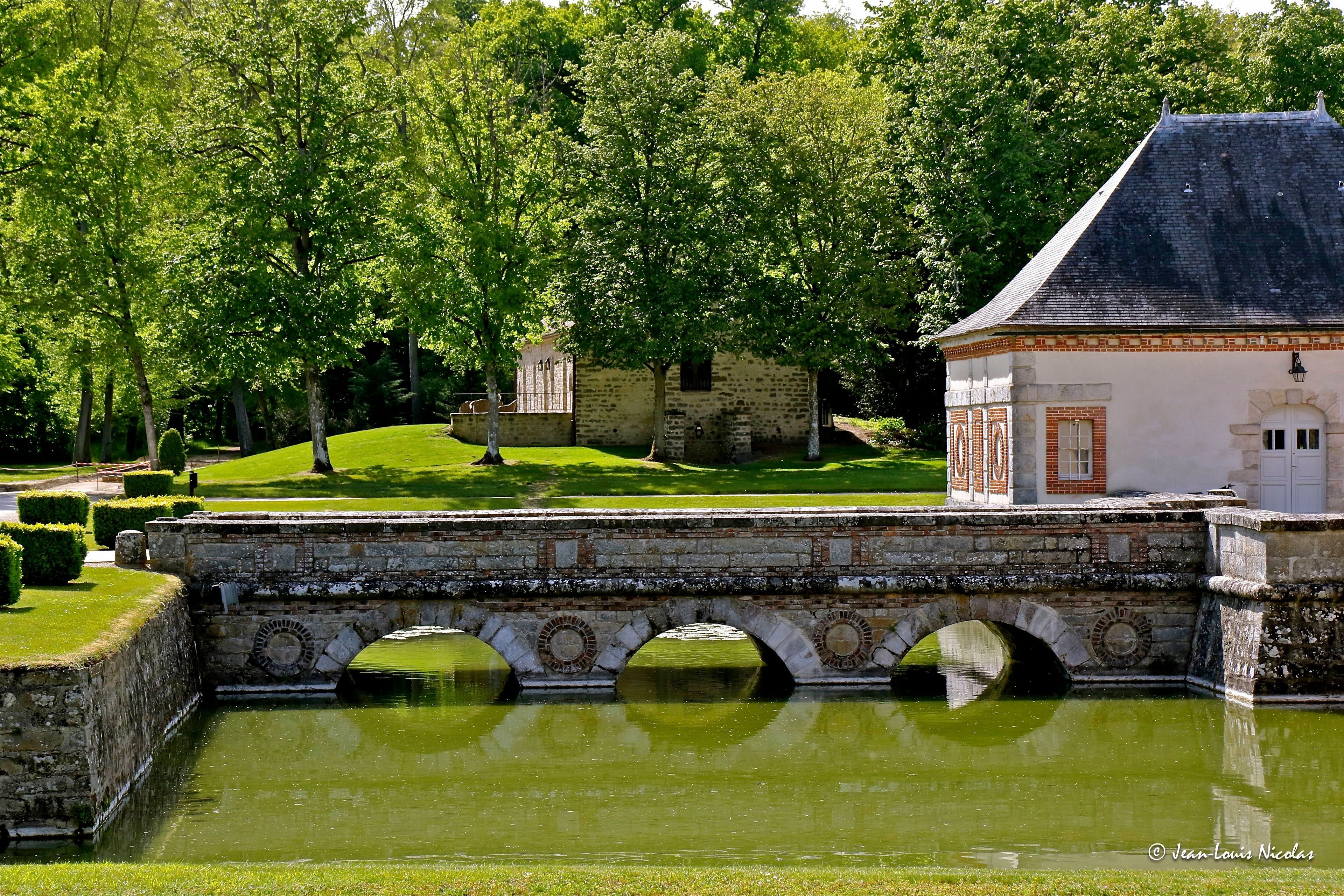 Un puente de piedra sobre una masa de agua tranquila, con un edificio de techo a la vista y un pequeño jardín al fondo.