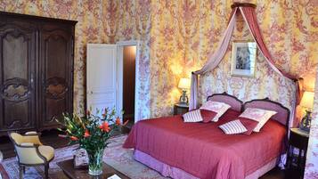 A bedroom with a large bed, a wooden wardrobe, a chair, a table with a vase of flowers, and a picture frame on the wall.