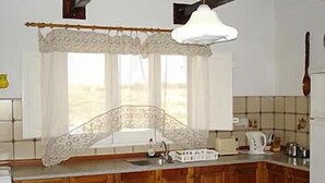A kitchen with wooden cabinets, a white countertop, a hanging light fixture, and a window with a lace curtain. - Villa Veduta (Ghajnsielem)