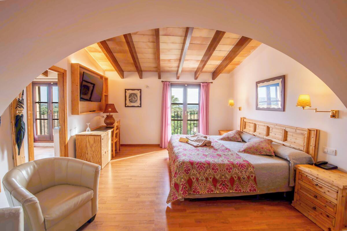 A bedroom with a wooden ceiling, a bed with a floral bedspread, a nightstand, a chair, and a window with curtains.