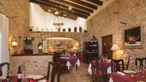 Un coin repas rustique avec des murs en pierre, des poutres en bois et un lustre. Les tables sont dressées avec des verres à vin et des assiettes.