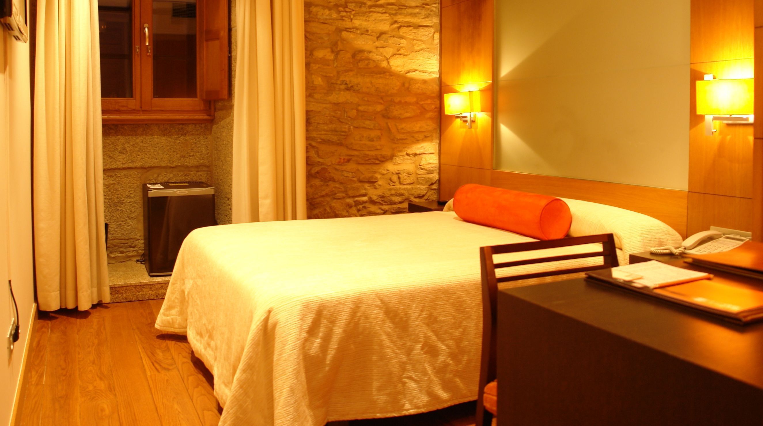 A hotel room with a bed, a desk with a telephone, and a stone wall.
