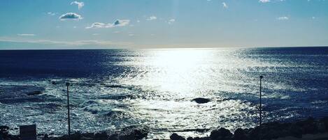 A view of the ocean with the sun reflecting off the water. There are rocks and a parking area in the foreground.