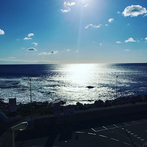 A view of the ocean with the sun reflecting off the water. There are rocks and a parking area in the foreground. - Il Nuovo Gabbiano (Dorgali)