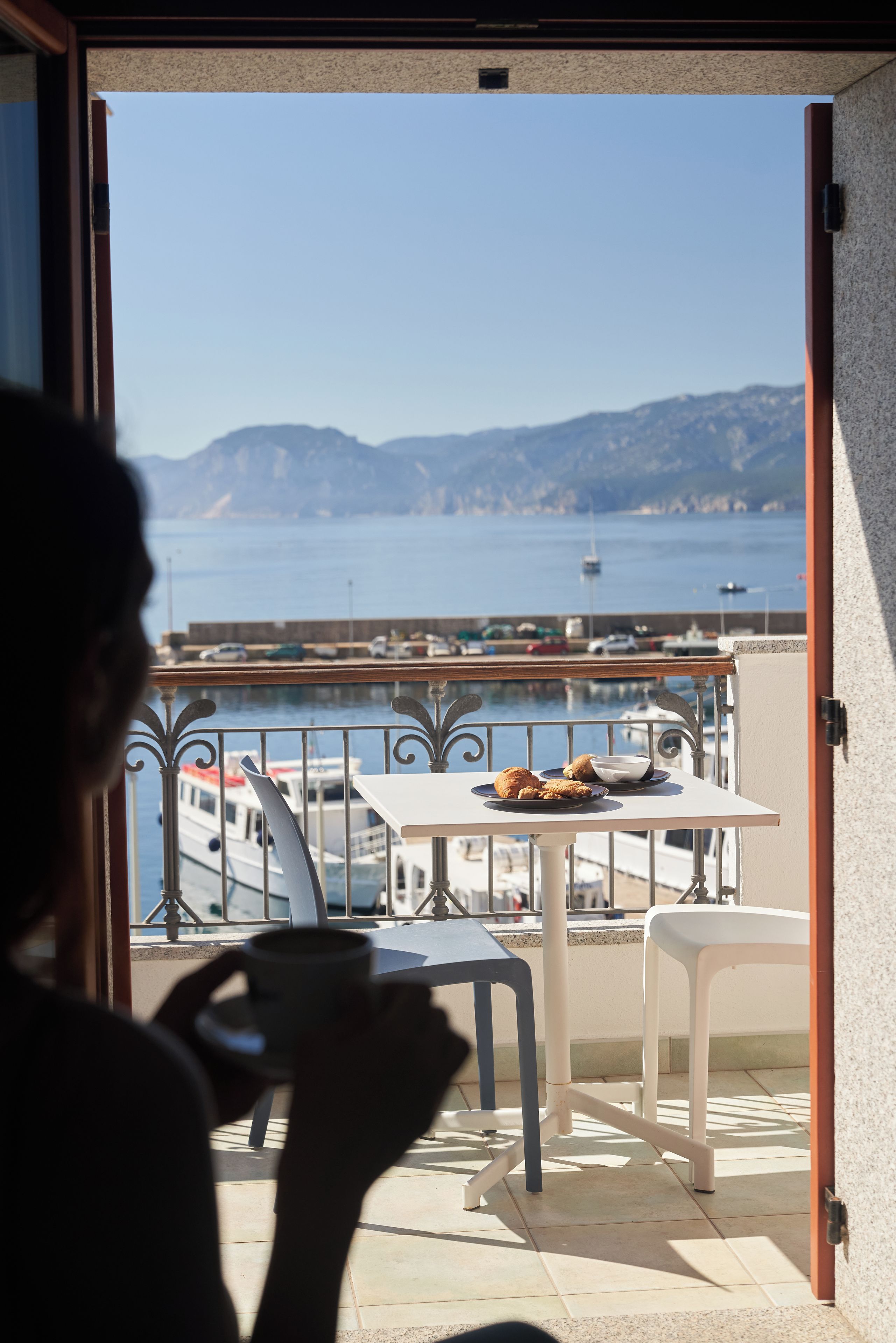 A balcony with a table set for breakfast, overlooking a marina with boats and a clear view of the sea and distant hills.