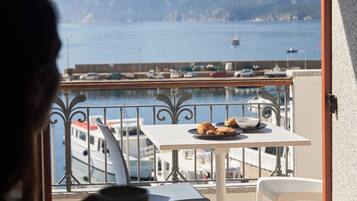 A balcony with a table set for breakfast, overlooking a marina with boats and a clear view of the sea and distant hills.
