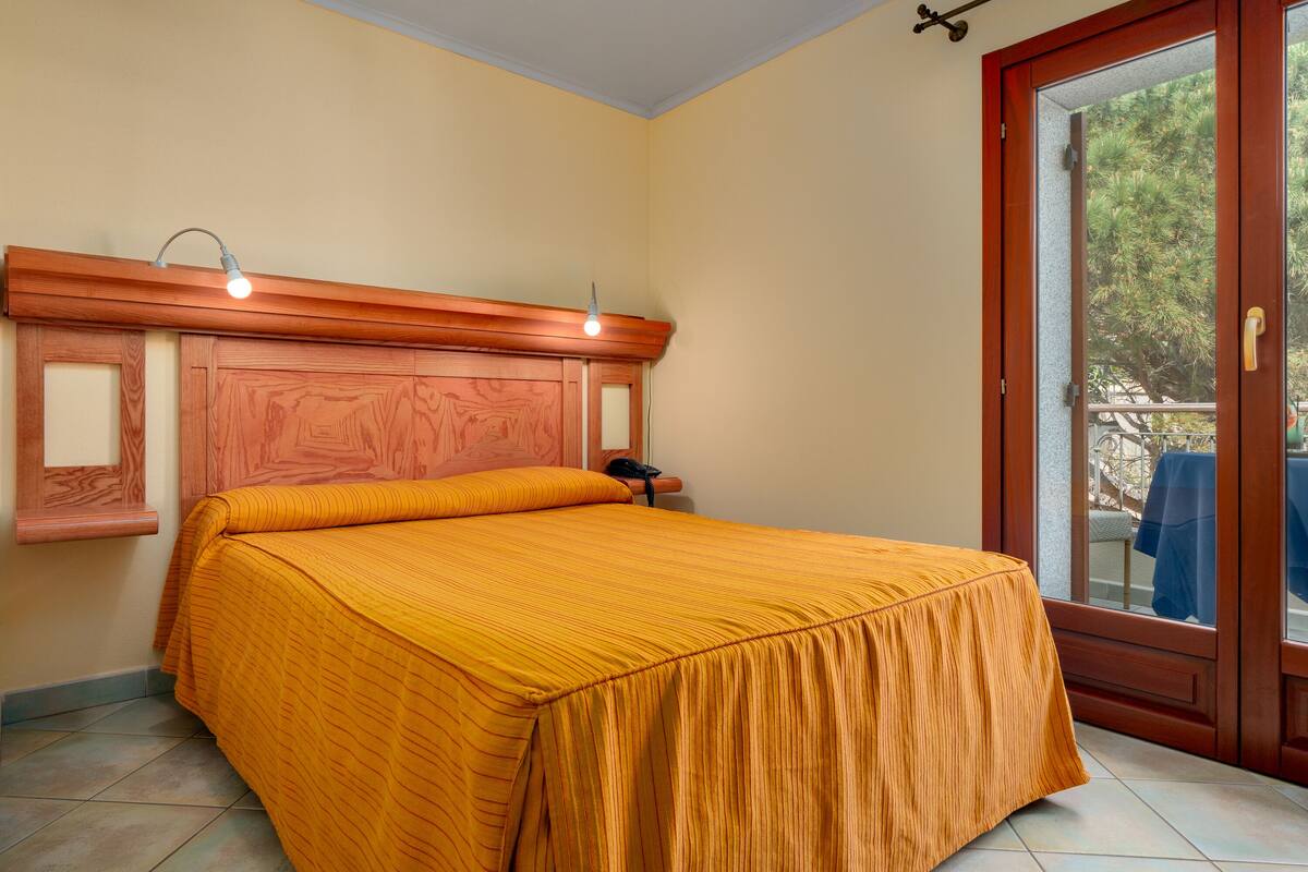 A bedroom with a wooden headboard, a bed with an orange bedspread, a bedside table with a lamp, and a sliding glass door leading to an outdoor area.