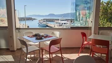 Zona pranzo con tavolo e sedie, vista su barche in marina e una porta vetrata che riflette la scena.