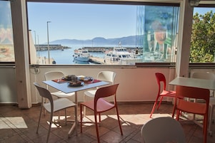 Un comedor con mesa y sillas, vista a barcos en un puerto deportivo, y un vidrio que refleja la escena.