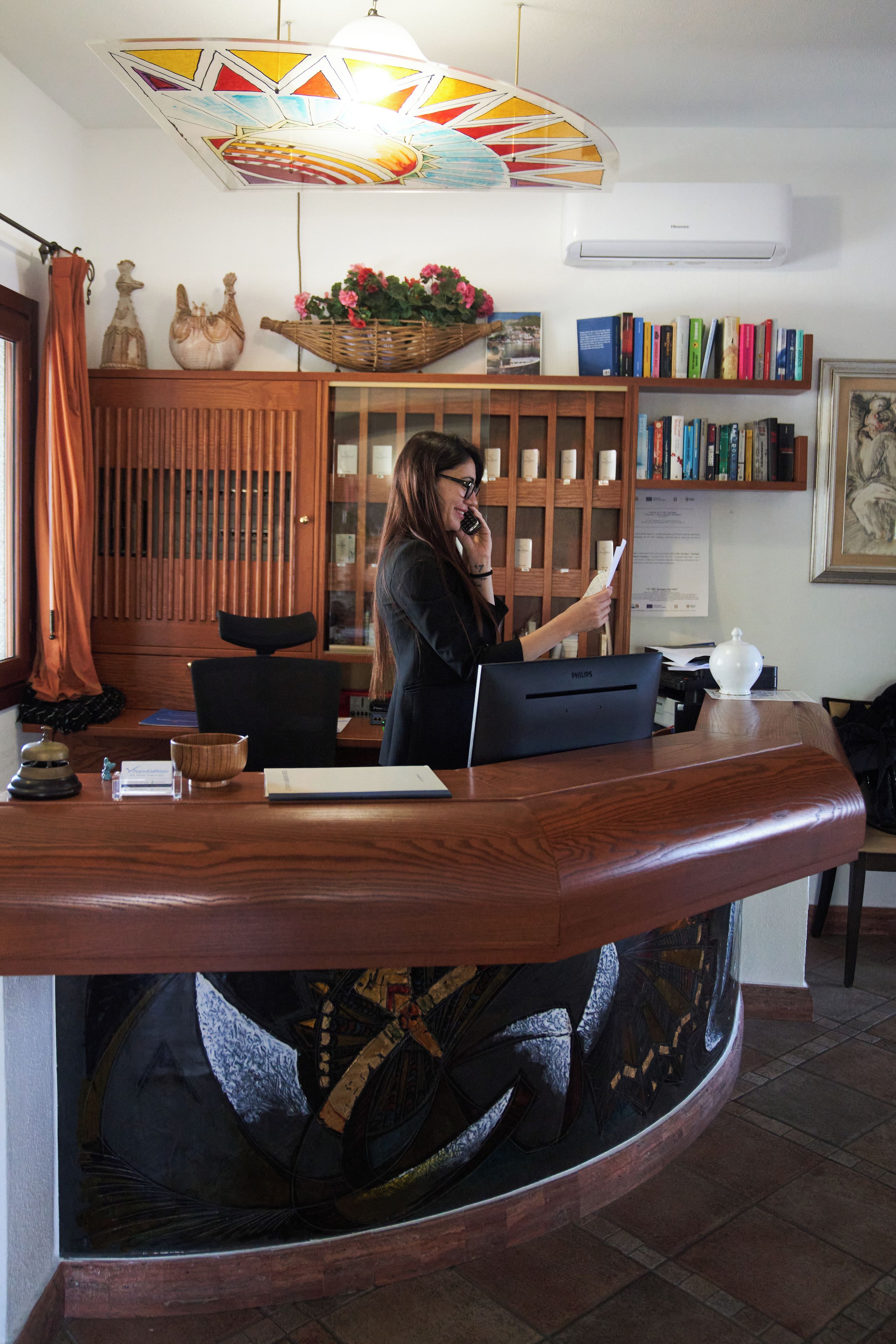 Una recepción con una mujer detrás, sosteniendo un documento. Se ve una estantería con varios libros y un cuadro en la pared.