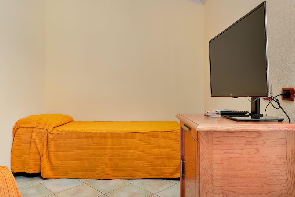 A single bed with a yellow bedspread, a wooden desk with a computer monitor, and a tiled floor.