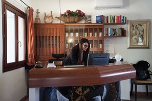 A reception desk with a computer, a telephone, and a small table with a bowl and a cup. There are bookshelves with books and a framed picture on the wall. - Il Nuovo Gabbiano (Dorgali)