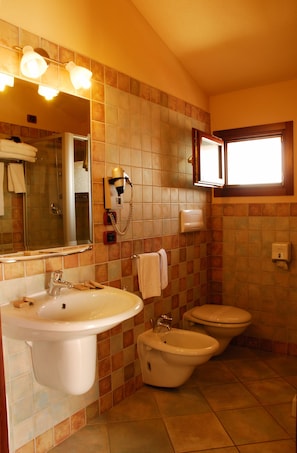 A bathroom with a sink, bidet, and toilet, tiled in a warm color scheme, with a mirror and lights above the sink. - Il Nuovo Gabbiano (Dorgali)