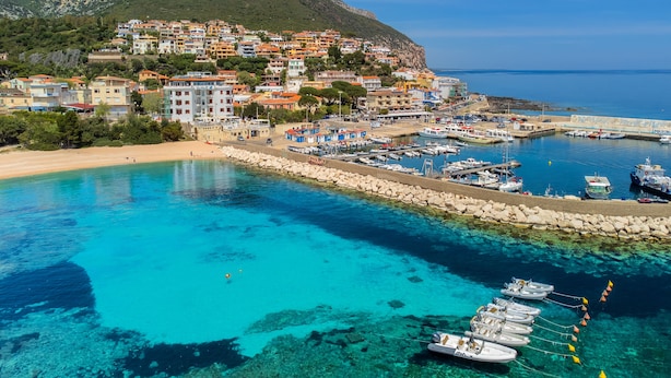 Un pueblo costero con un puerto deportivo, aguas azules cristalinas y edificios en una colina.