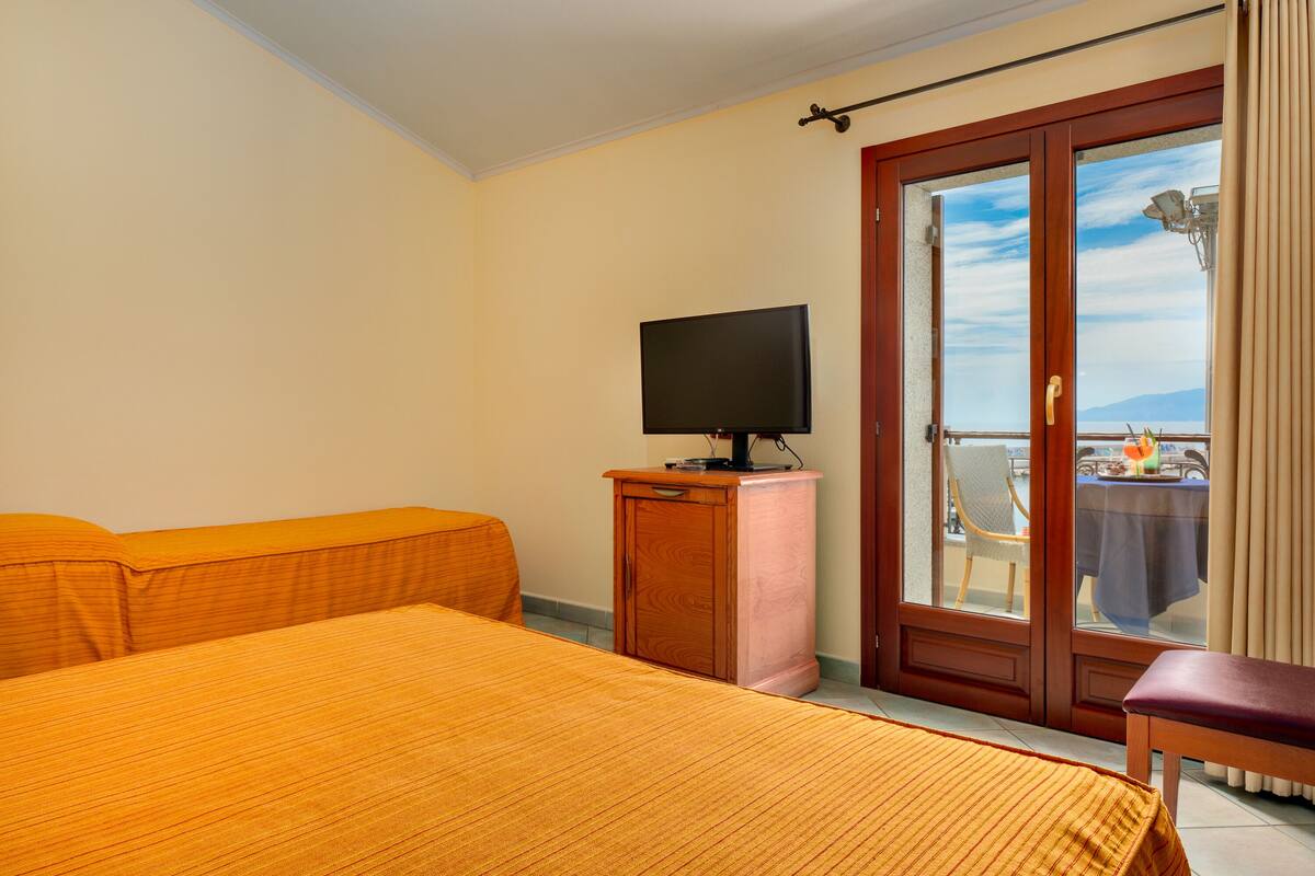 A hotel room with a bed, a television on a wooden cabinet, and a balcony with a table and chairs.