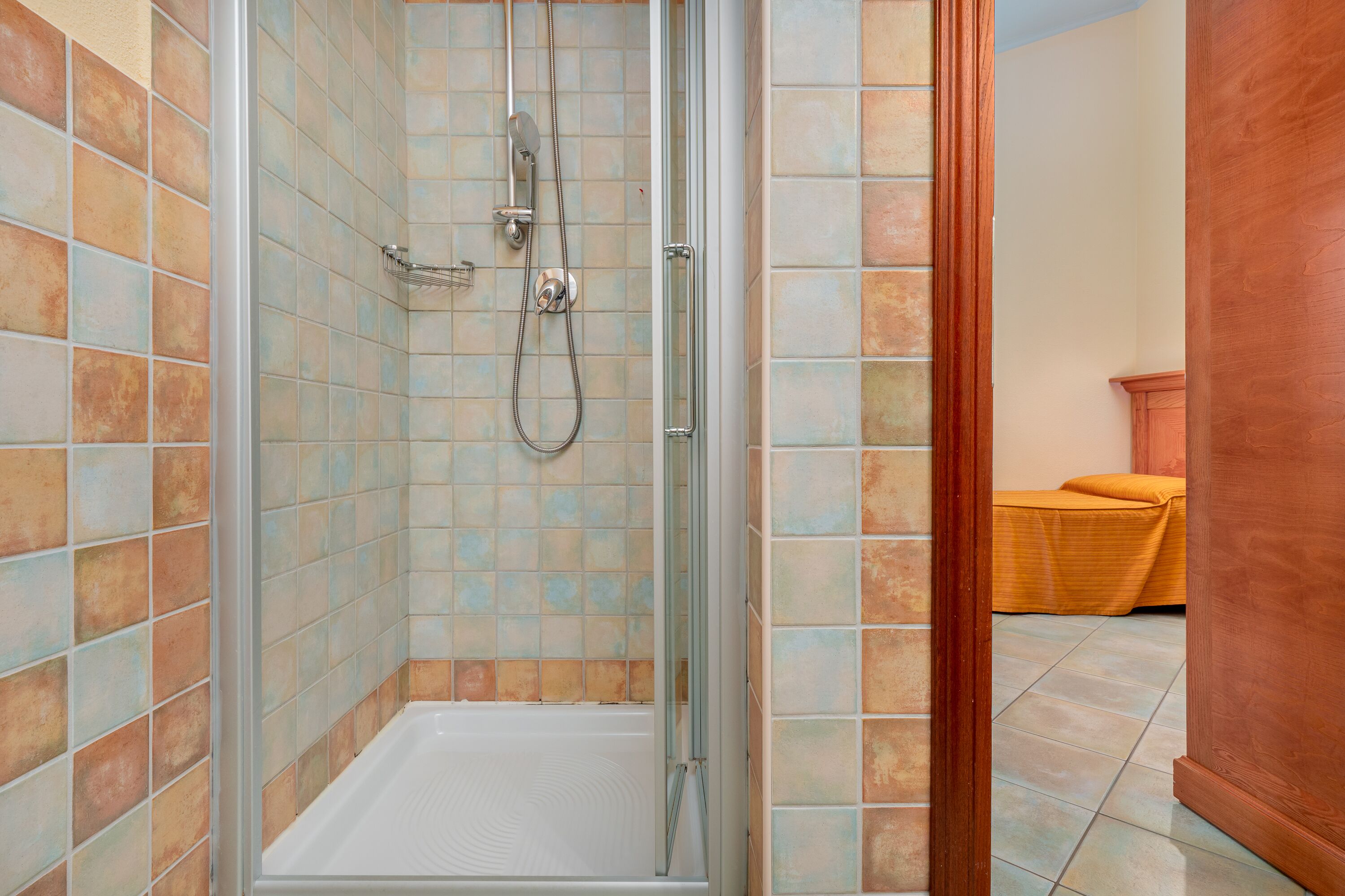 A shower area with a glass door, tiled walls, and a wooden bench in the background.