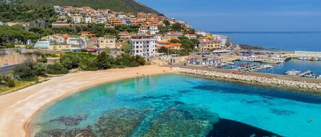 A coastal town with a clear beach, a marina, and buildings with red-tiled roofs.