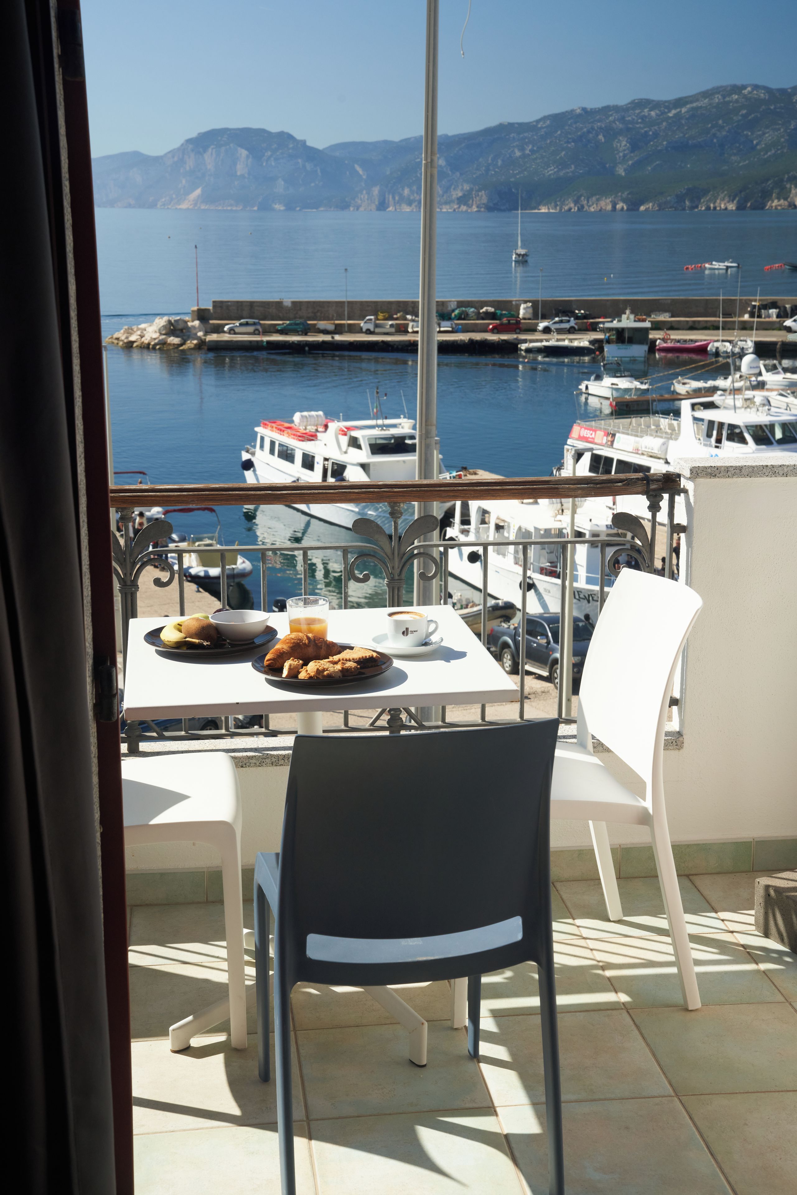 Un balcon avec une table dressée pour le petit-déjeuner, donnant sur une marina où sont amarrés des bateaux, et offrant une vue dégagée sur la mer et les montagnes lointaines.