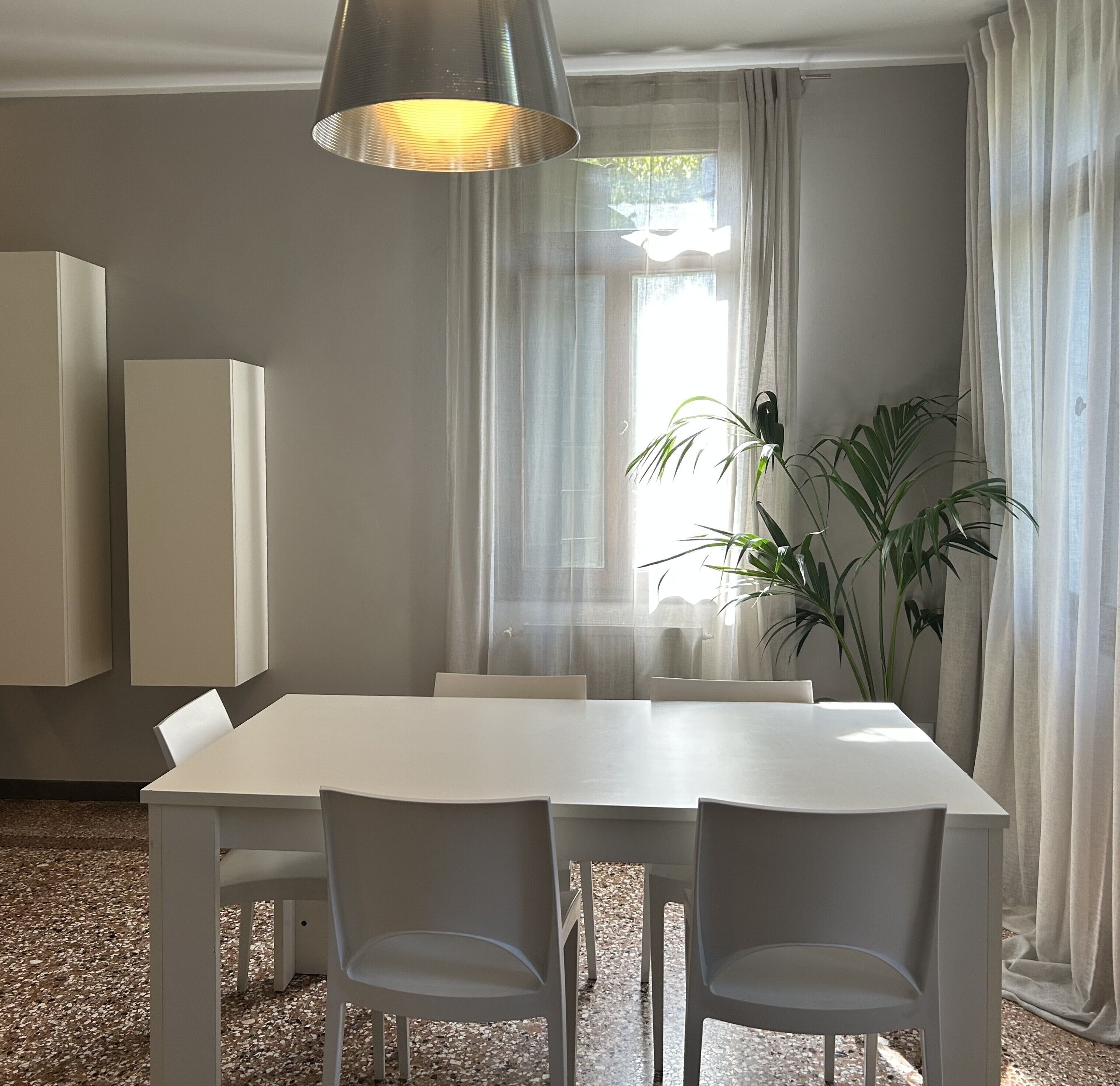 A modern dining area with a white table and chairs, a hanging light fixture, and a large window with sheer curtains.