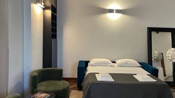 Una habitación de hotel con una cama, un sillón verde, una mesita con un libro y un espejo.