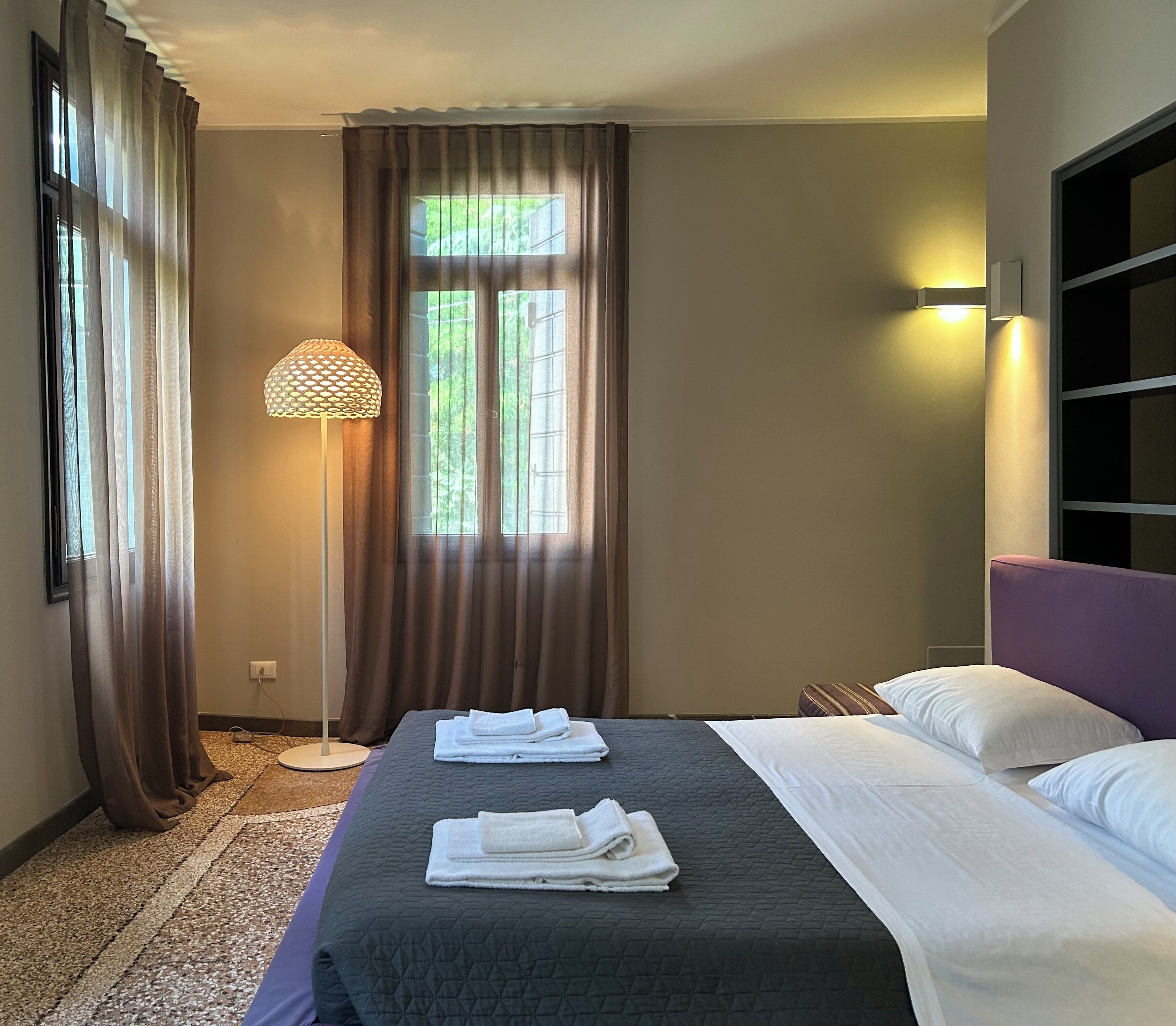 Quarto de hotel com cama, luminária de chão e janela com cortinas.