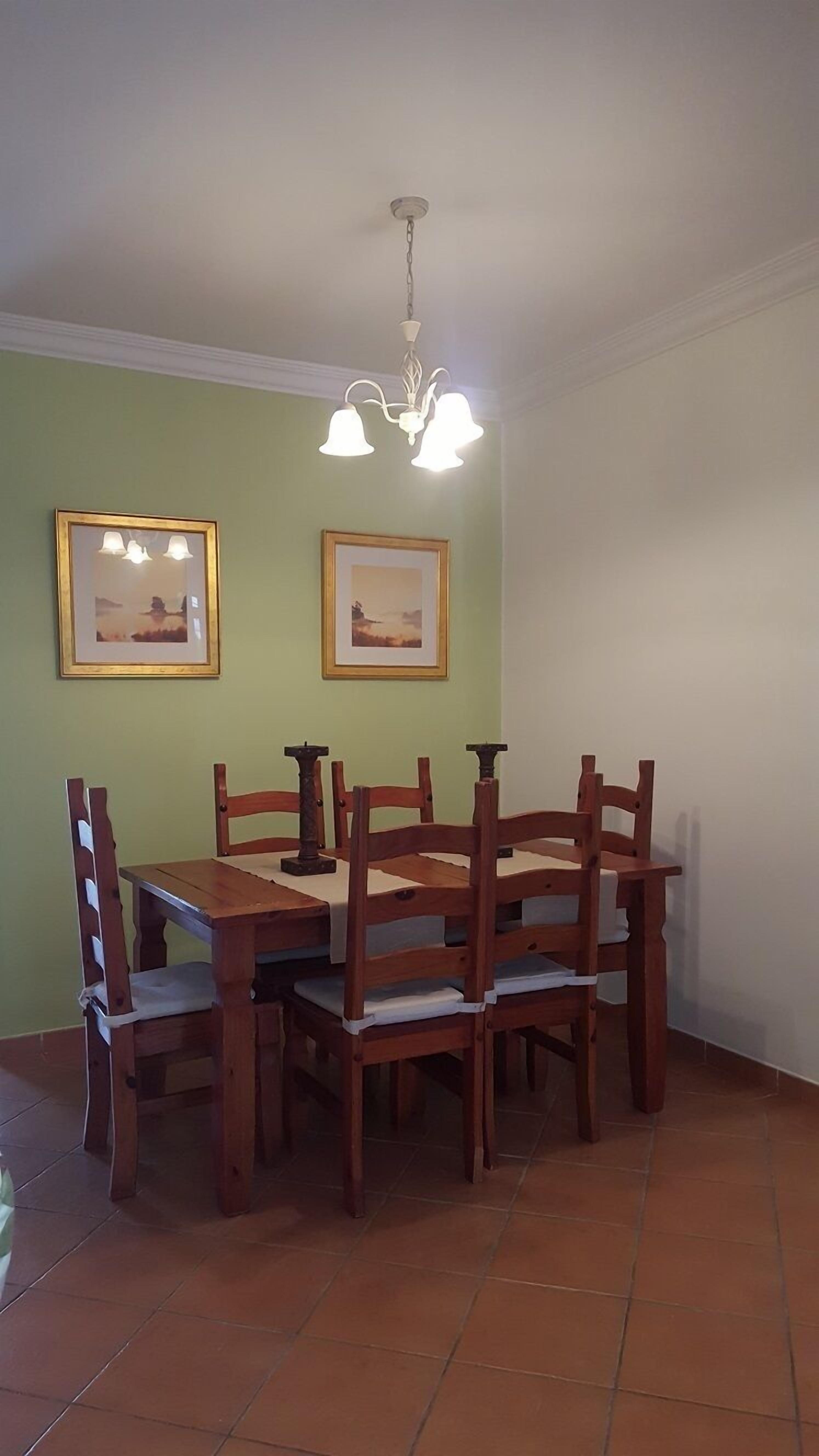 A dining area with a wooden table and chairs, two framed pictures on the wall, and a hanging light fixture.
