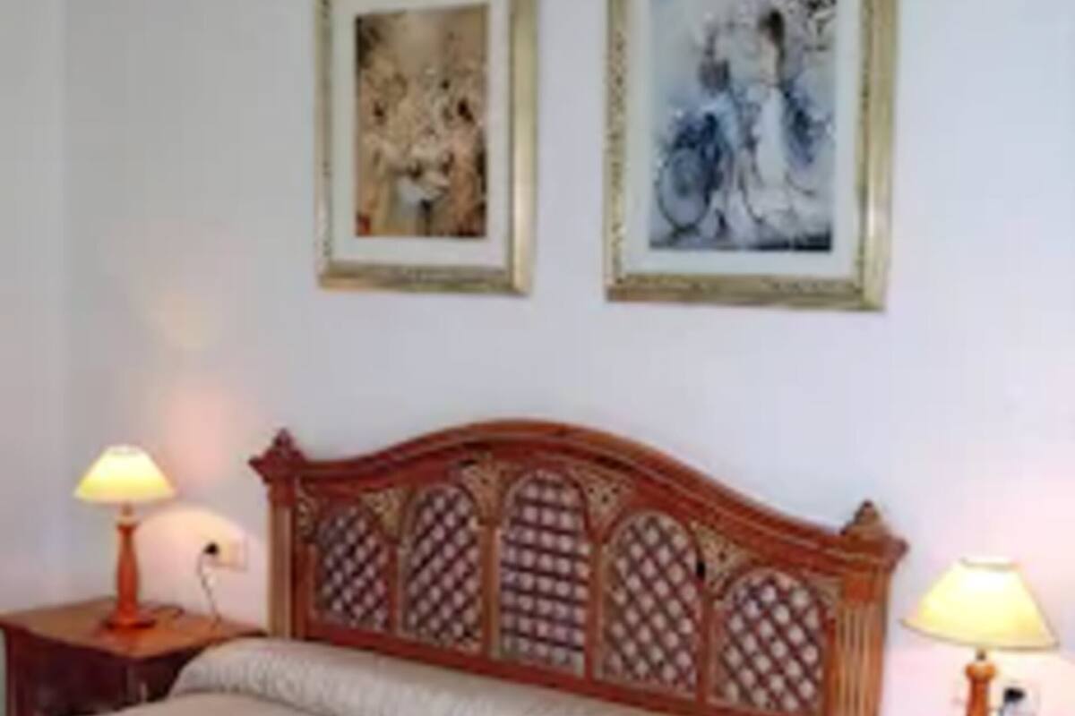 A bedroom with a wooden headboard, two bedside tables with lamps, a bed with a white bedspread, and two framed pictures on the wall.