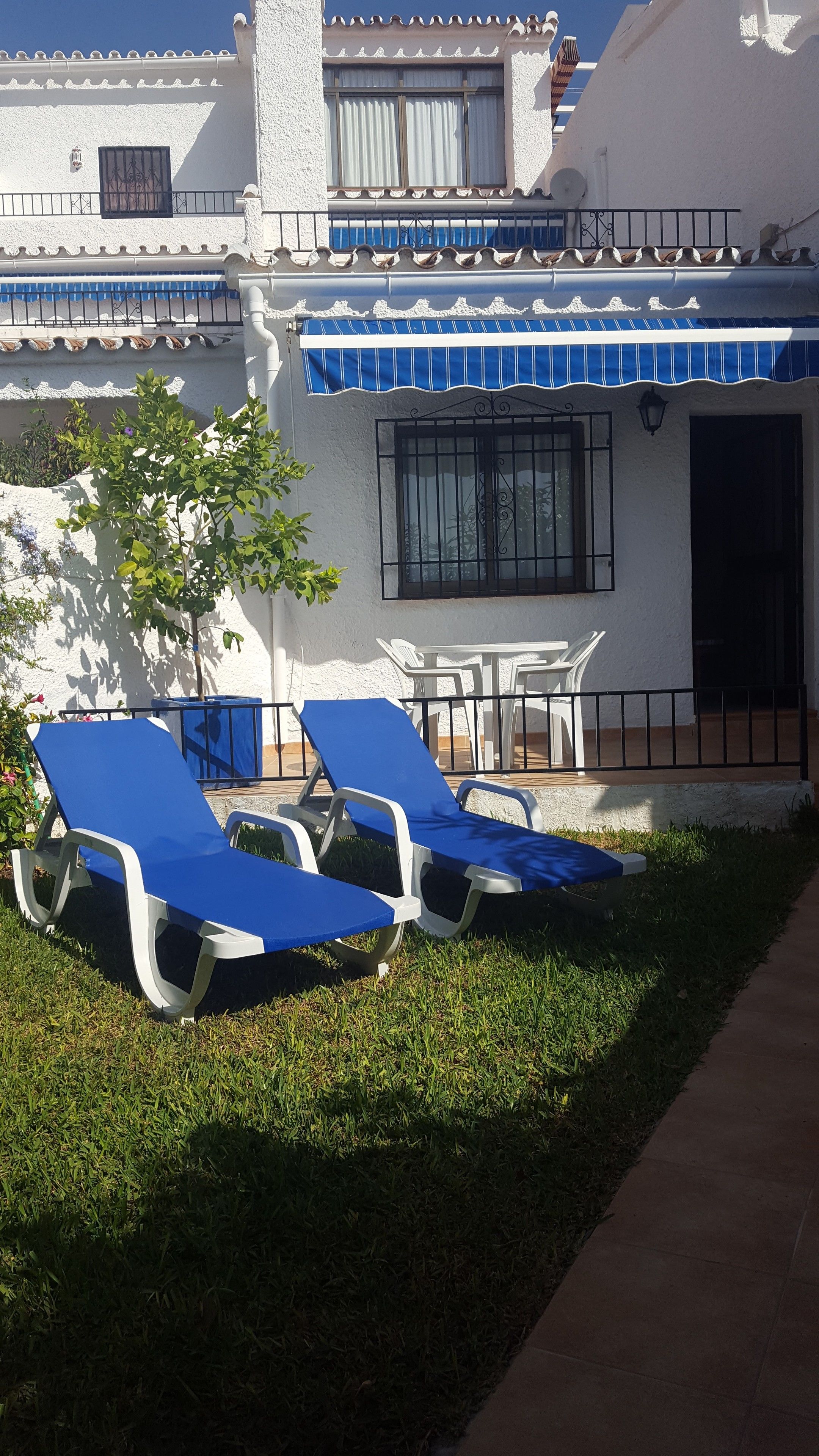 A building with a white exterior, blue-striped awning, and a balcony with a black railing. There are two white tables and four blue lounge chairs arranged outside.
