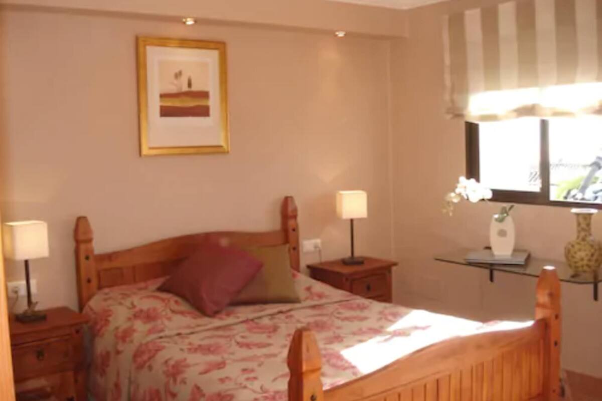 A bedroom with a wooden bed, two bedside tables, a nightstand, a framed picture on the wall, a window with curtains, and a vase with flowers on a small table.
