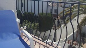 Uma varanda com uma cadeira azul, guarda-corpo de metal e vista para uma rua com edifícios e céu claro.