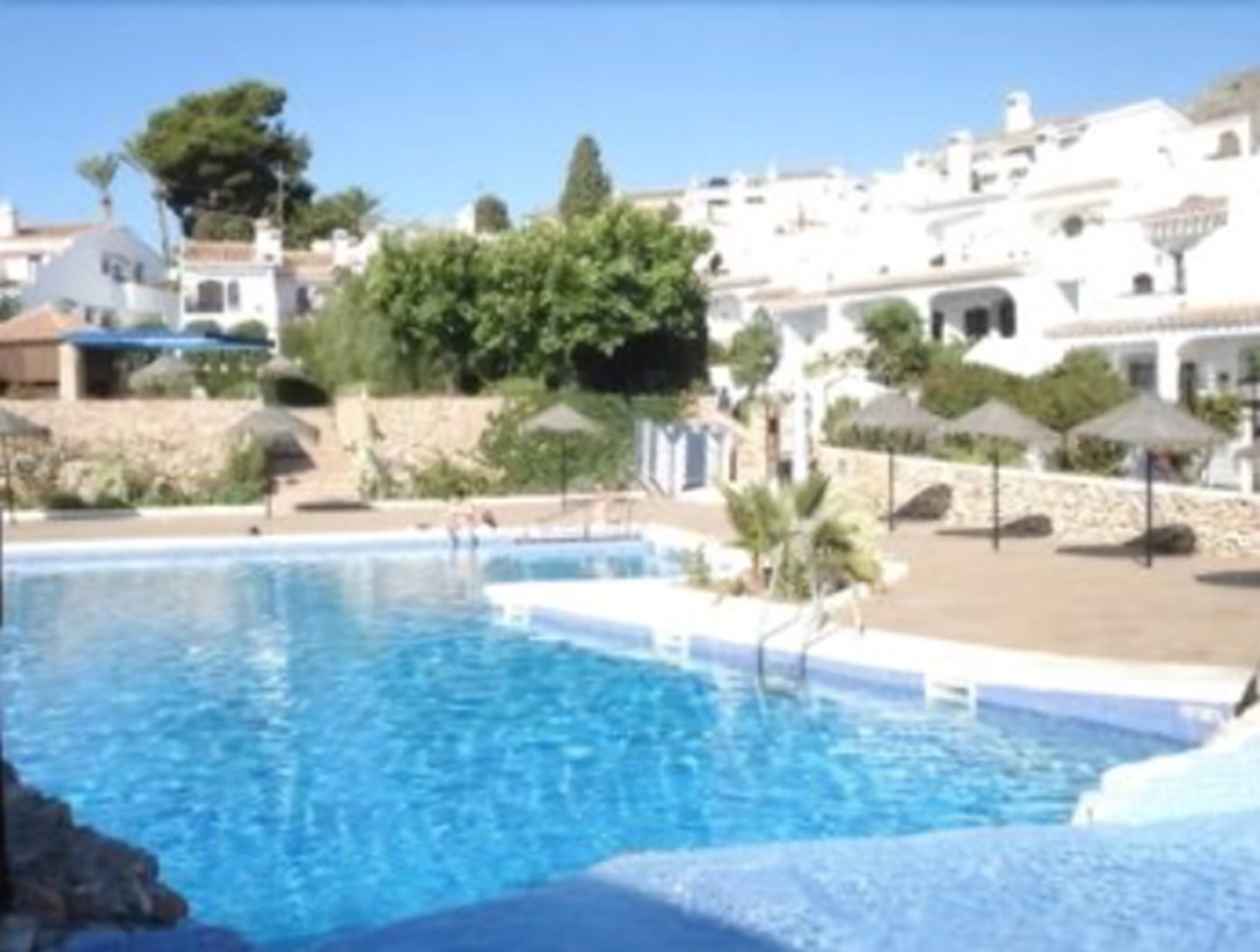 A swimming pool with clear blue water, surrounded by white buildings with stone walls and greenery.