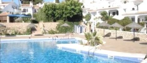 A swimming pool with clear blue water, surrounded by white buildings with stone walls and greenery.