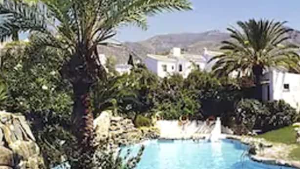 Piscina rodeada por pedras e palmeiras, com edifícios ao fundo.