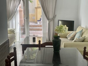 Ein Wohnzimmer mit einer Couch, einem Couchtisch und einem Fernseher. Durch eine Glastür ist der Außenbereich sichtbar.