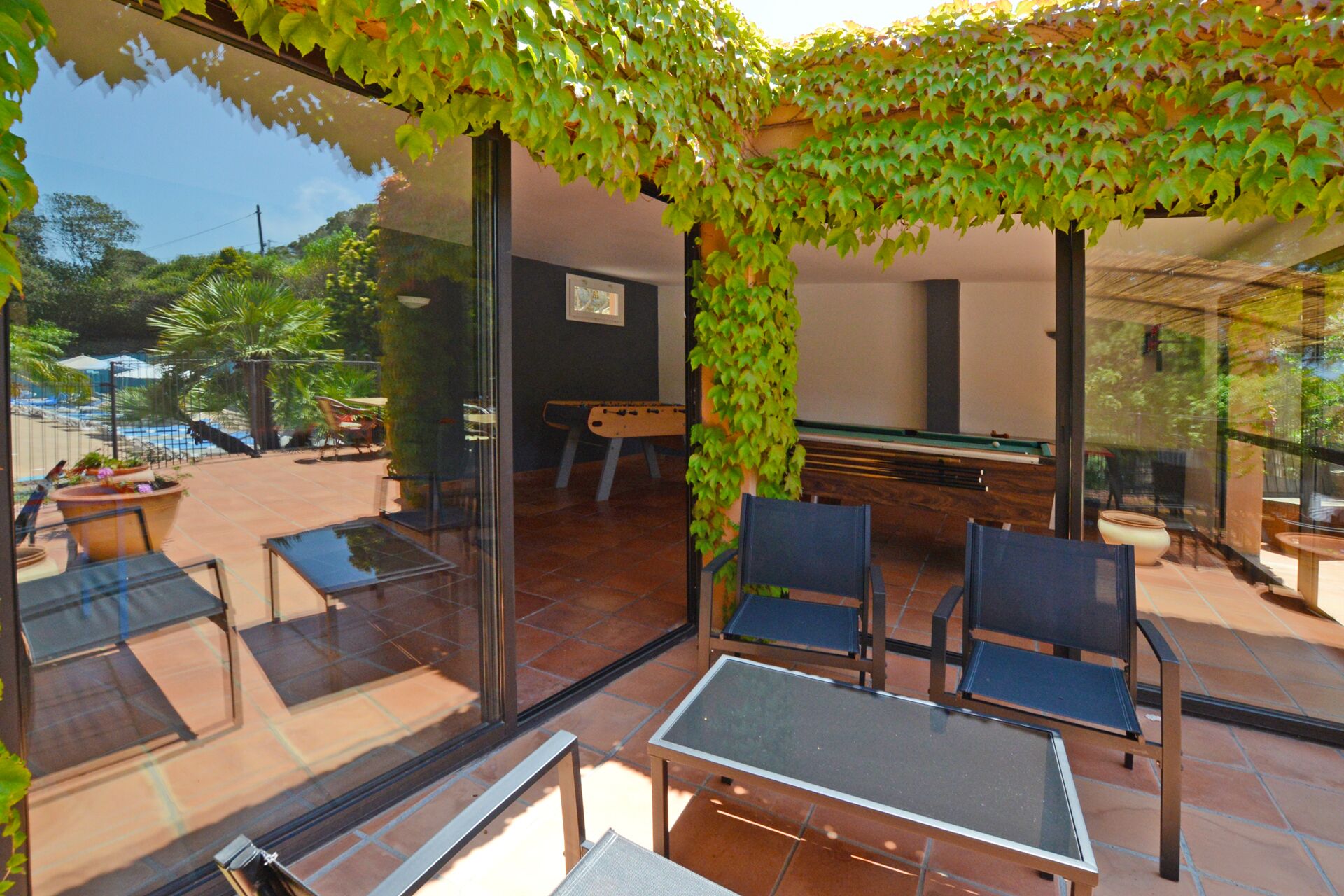 An outdoor patio area with a glass table and chairs, surrounded by a glass wall and a green vine-covered pergola.