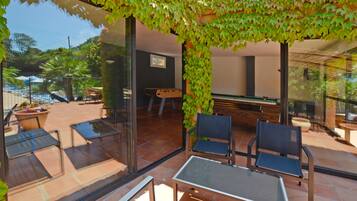 An outdoor patio area with a glass table and chairs, surrounded by a glass wall and a green vine-covered pergola.