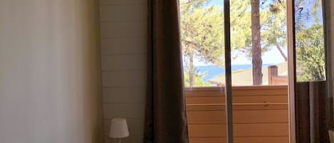 Un dormitorio con cama, una mesita de noche con lámpara, ventana con cortinas y vistas a árboles y un edificio.