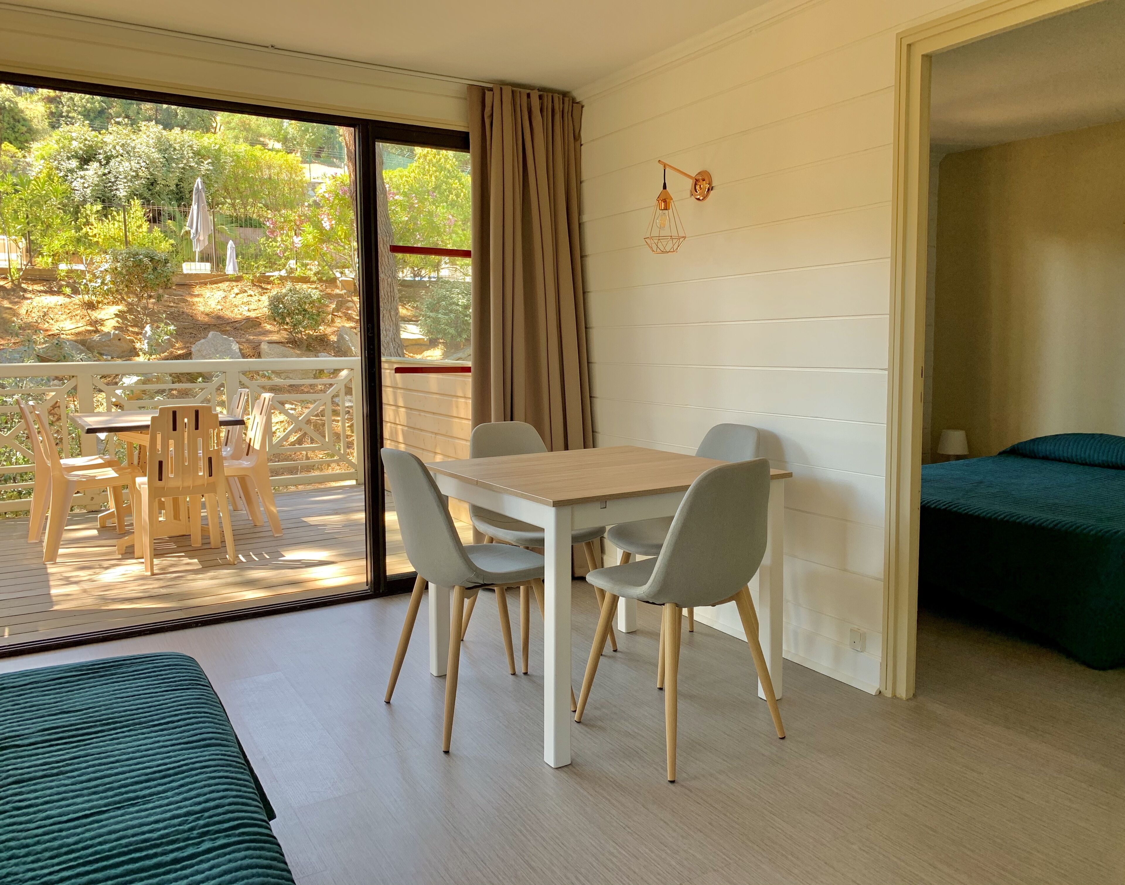 A room with a wooden table and chairs, a bed, and a view of a garden through a sliding door.
