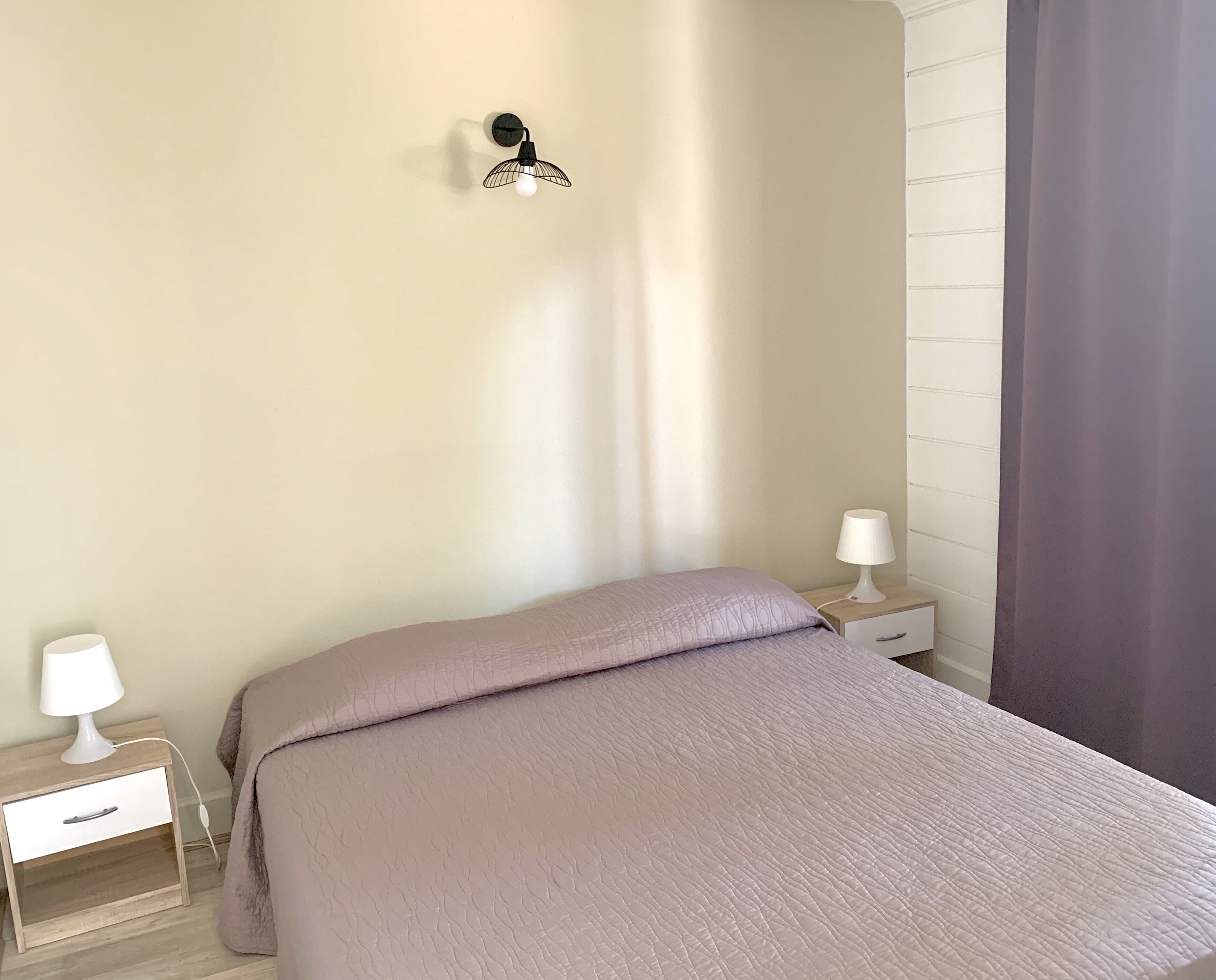 A bedroom with a bed, two bedside tables with lamps, a wall-mounted light fixture, and a window with a curtain.